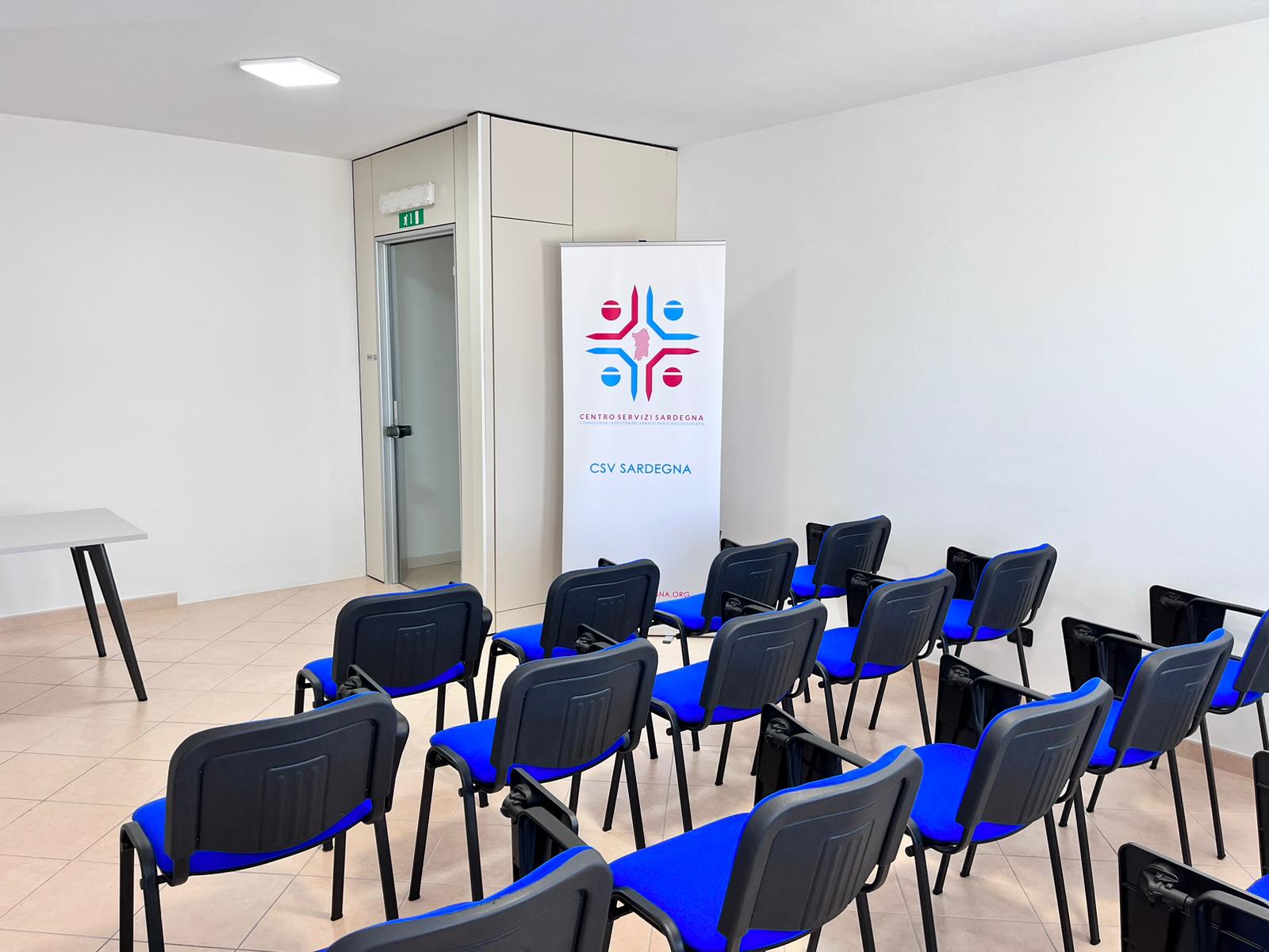 Sala conferenze con sedie blu e tavolo, vicino a un cartellone con logo colorato.